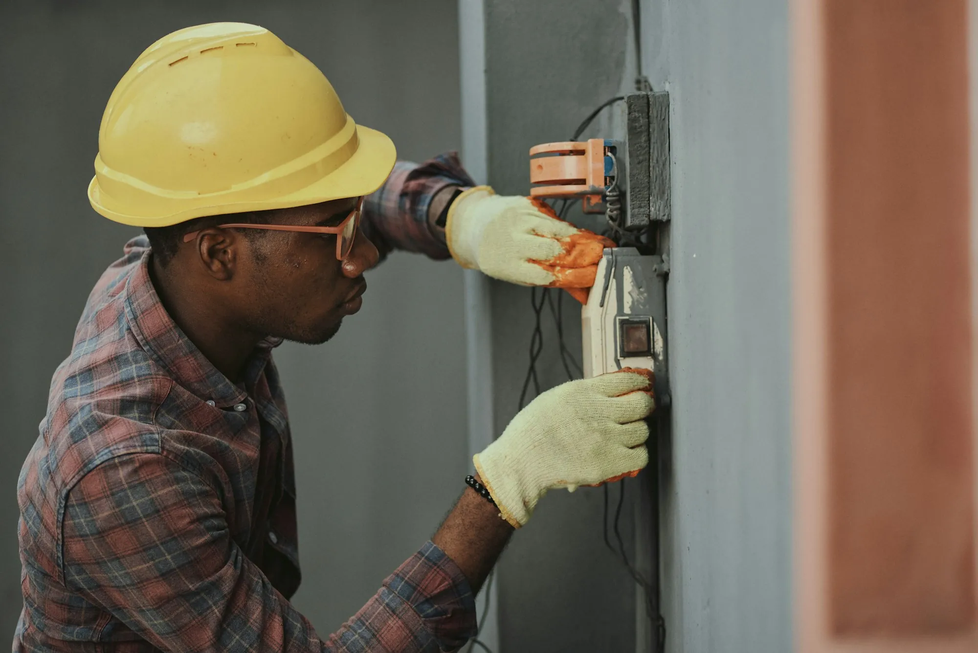 Tradesman in a yellow helmet wiring a gray meter box on an exterior wall